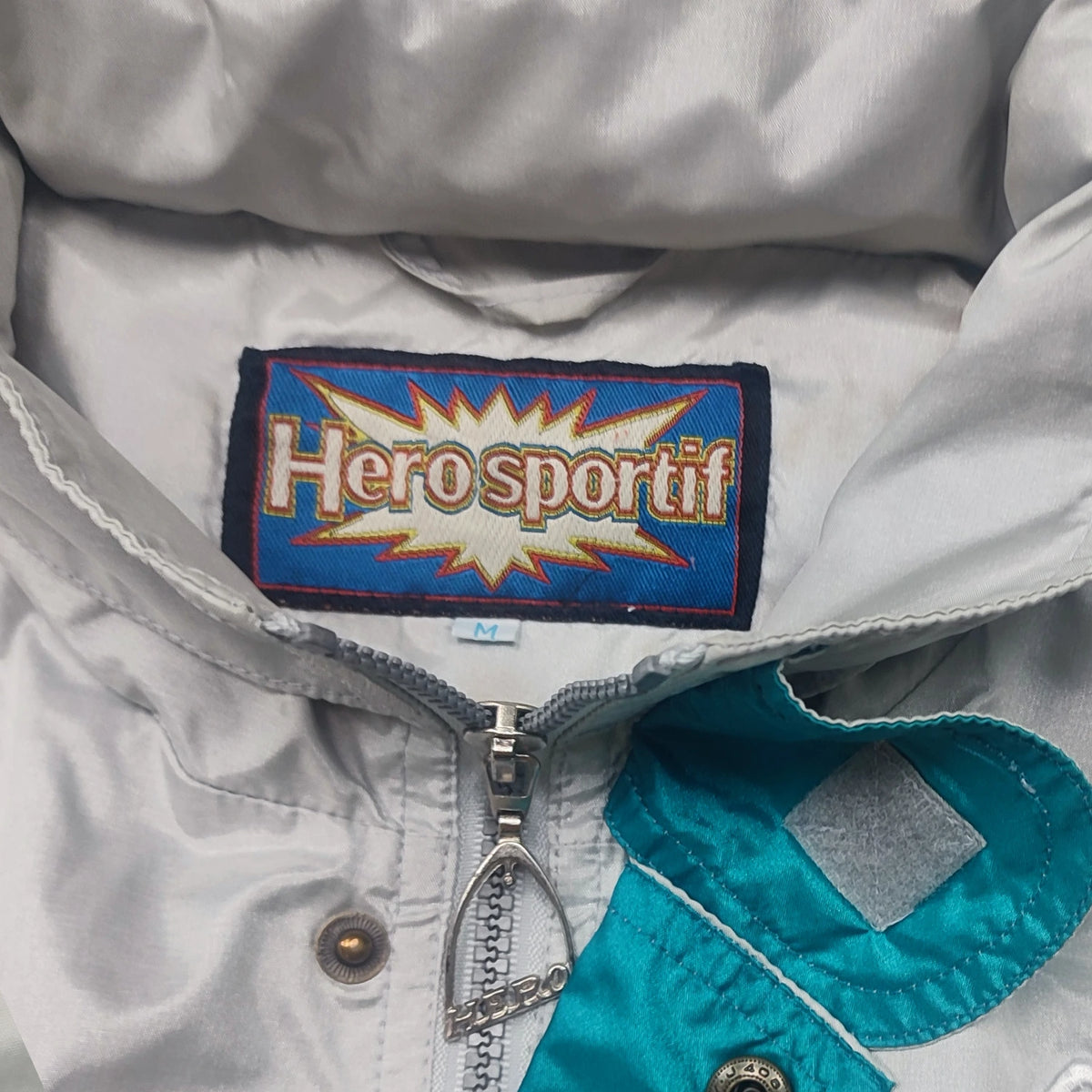 Herosportif-Etikett mit Größe M im hellgrauen Kragen neben türkiser Blende, Reißverschluss und Druckknopf.
